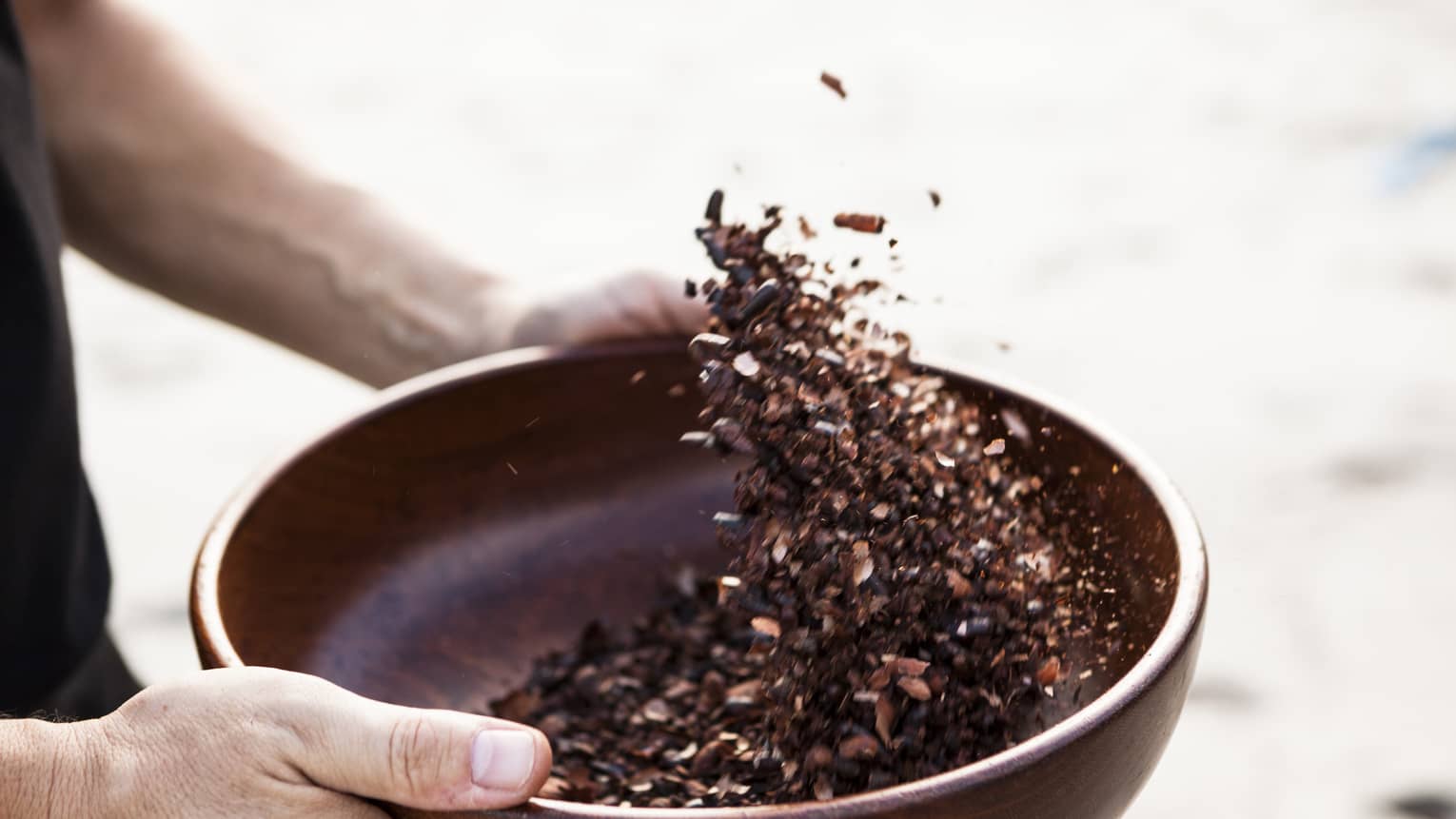 Close-up of hands holding dark wood bowl, shaking crushed cocoa