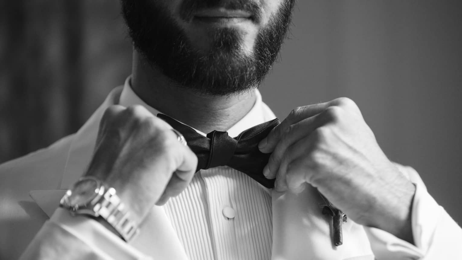 A black and white of a man in a white tuxedo tightening his bowtie
