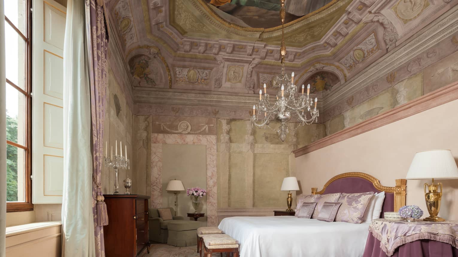 Frescoed Executive Suite bed with purple velvet headboard, nightstand with cloth under chandelier, vaulted ceiling