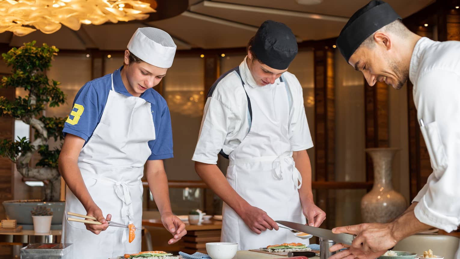 Two teenagers wearing aprons and chef hats learning to make sushi with a sushi chef