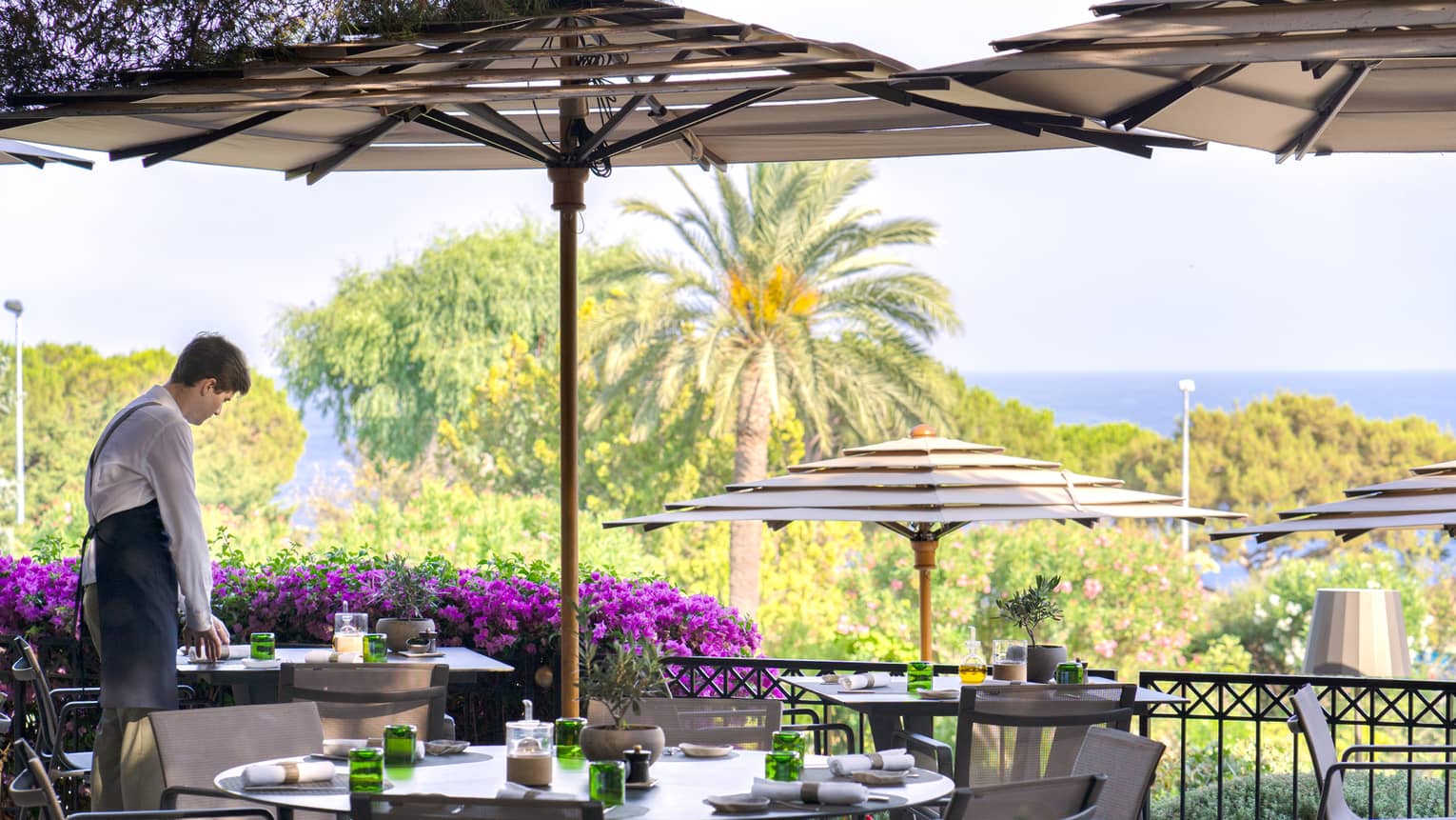 Server arranging napkin on La Véranda terrace under umbrella, palm tree and sea view