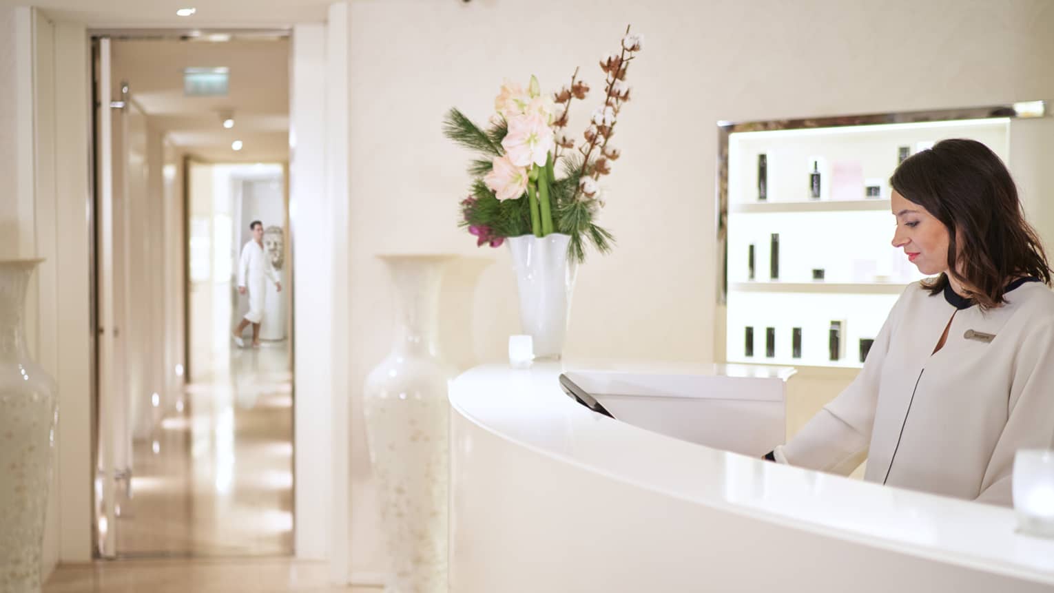 Staff member in white jacket standing behind curved white reception desk with flower vase, view into spa hallway