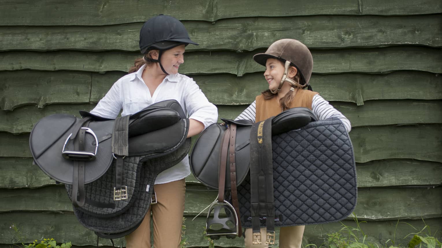 Two children in horseback-riding helmets smile as they carry saddles and pads against a backdrop of dark green barnboard.