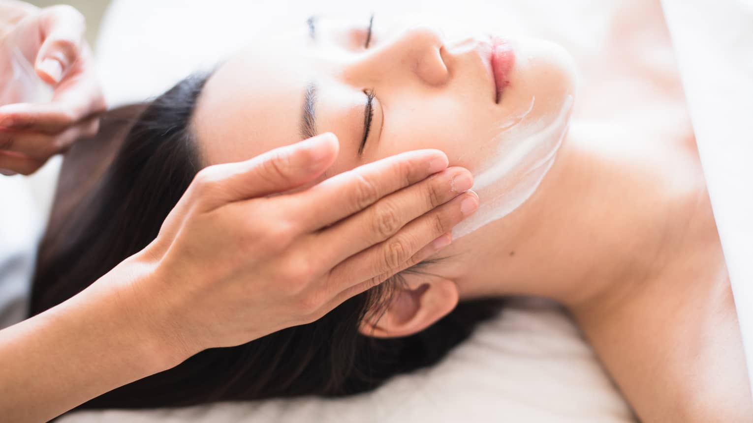 Close-up of spa attendant's hands rubbing white cream on woman's face as she lies on massage table