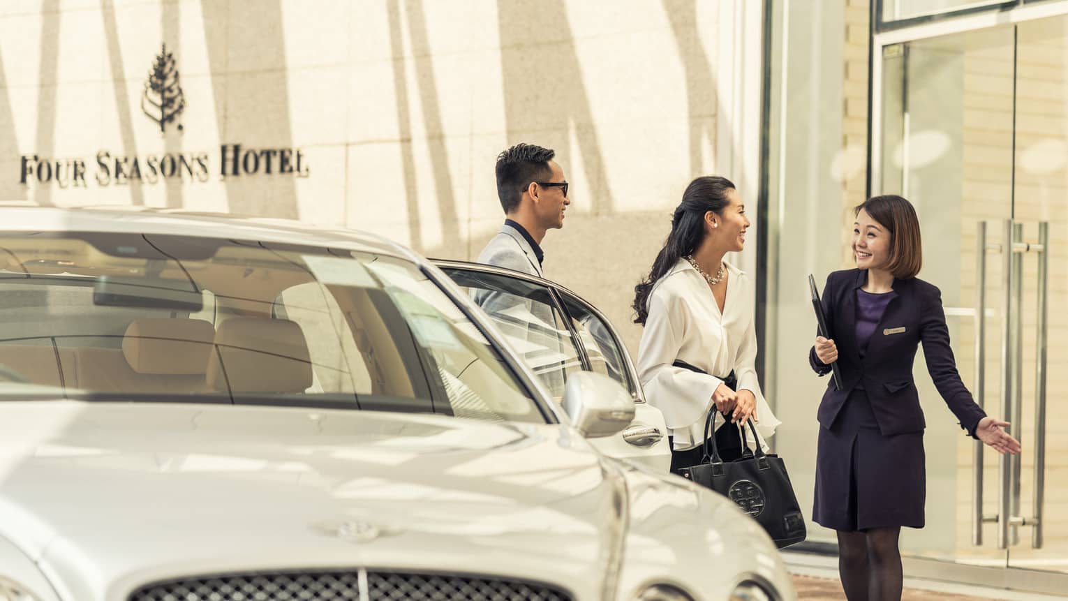 Hotel staff greets couple in front of luxury car at Four Seasons Hotel Hong Kong entrance