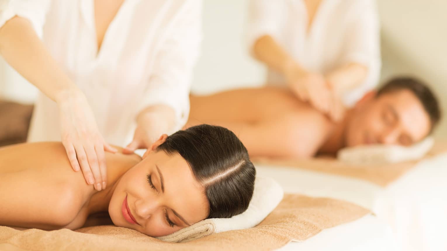 Woman and man lie side-by-side on massage tables as spa staff massage their bare shoulders