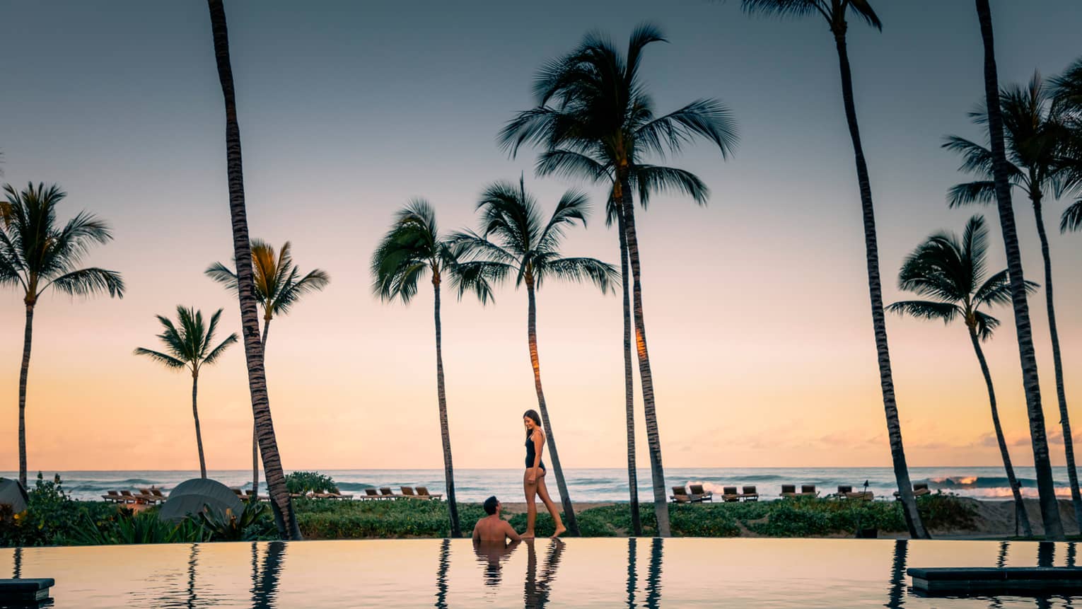 Woman in swimsuit walks on edge of pool past wading man, tall palm trees at sunset