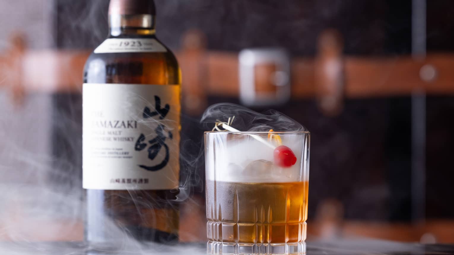 Bottle of Japanese whisky beside half-full cut-crystal glass with red cherry garnish, dry ice mist swirling from the glass.