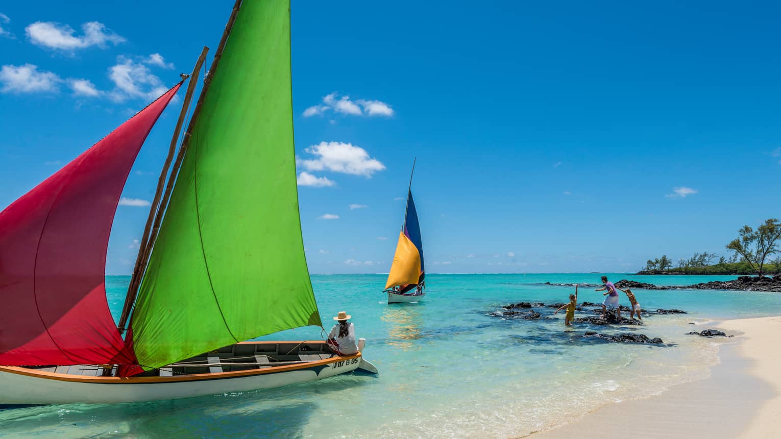 Family plays on beach, rocks in front of two sailboats with colourful sails