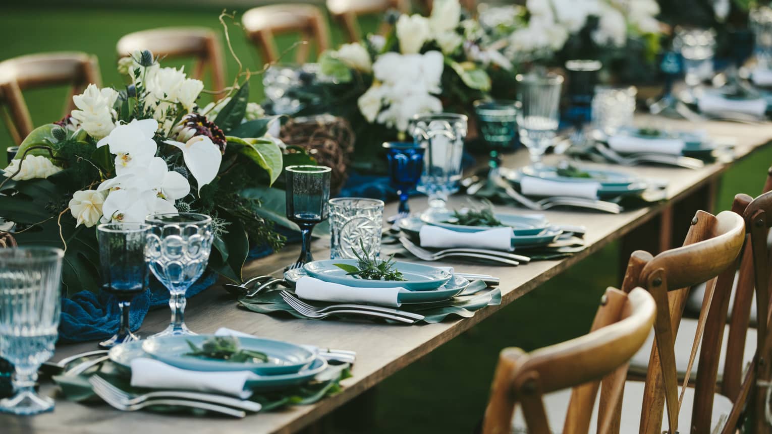 A close up of a long wooden table set with blue plates, eclectic blue and clear glassware, white napkins and green and white floral arrangements all the way down the center of the table