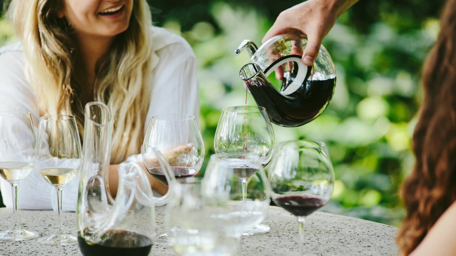 Smiling women at outdoor table, hotel staff pours red wine from artistic glass carafe