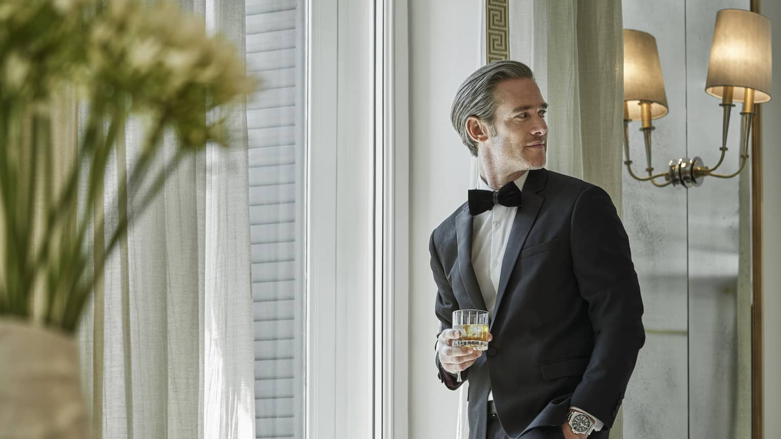 A man in a black tuxedo holds a glass of whiskey while waiting in a white doorway.