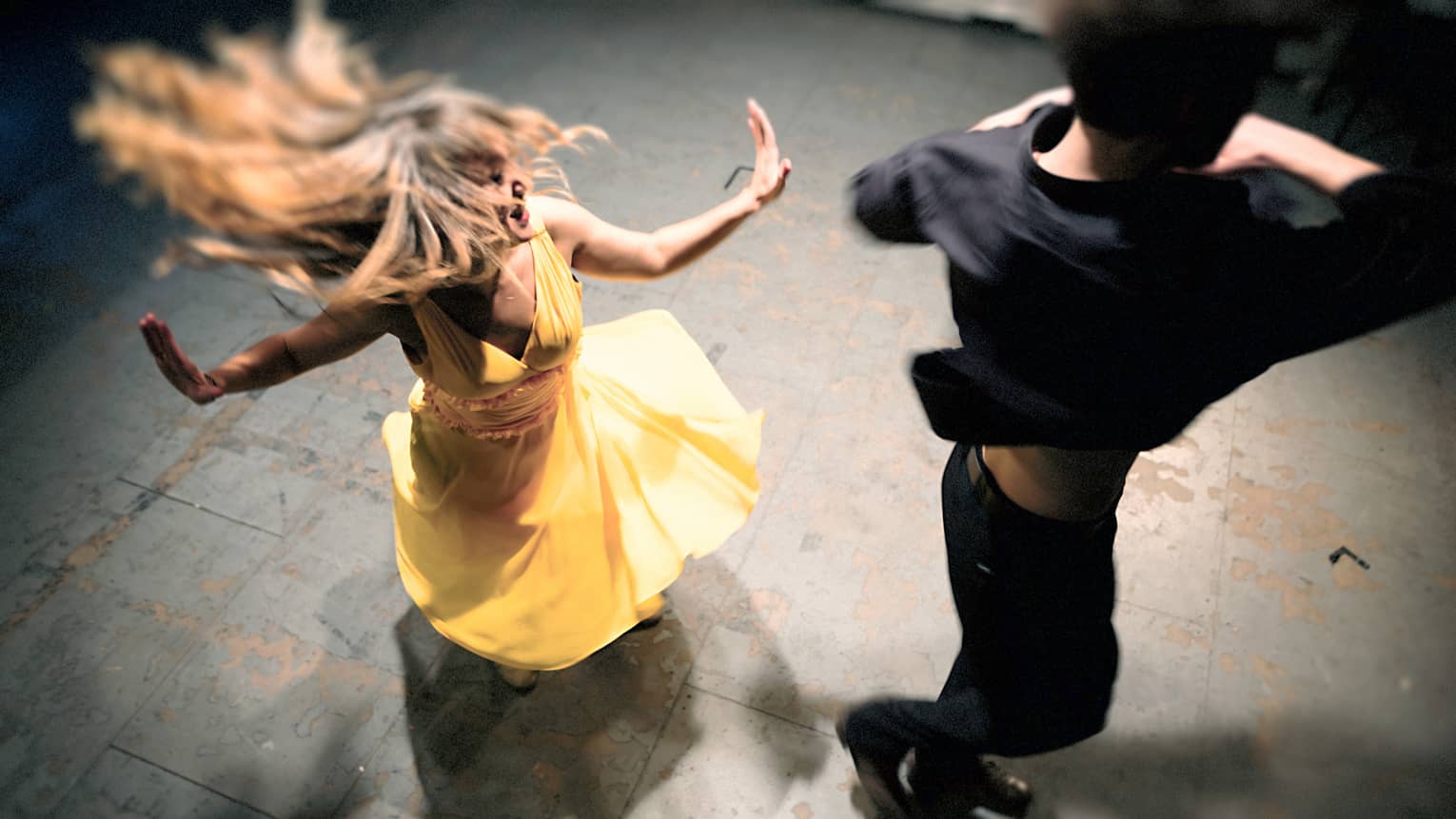 Woman in a yellow dress is spinning on the dance floor as a man wearing all black dances next to her