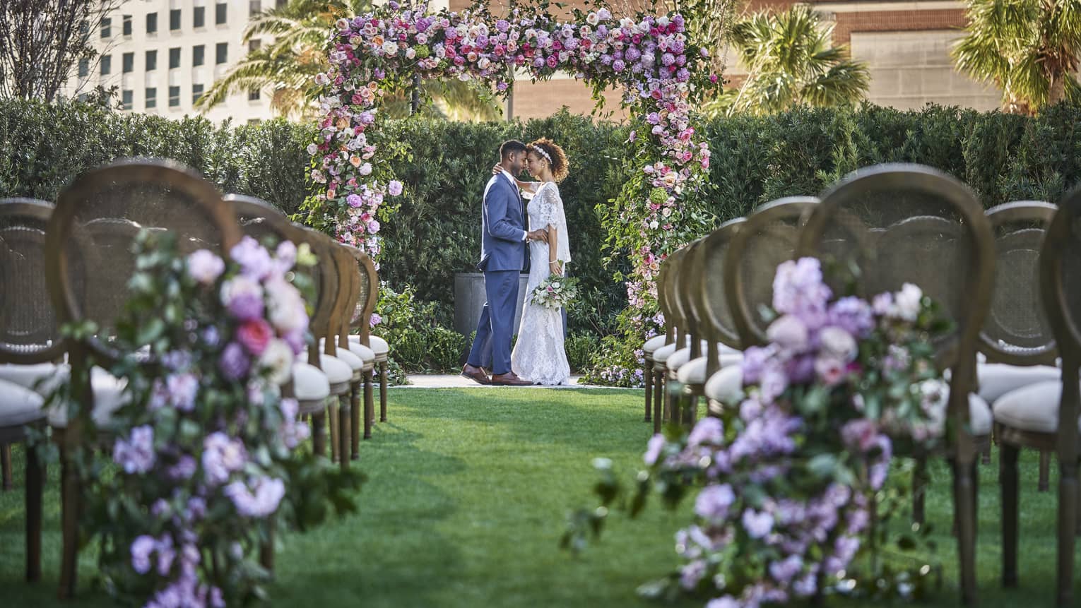 A bride and groom under an arch of flowers with chairs lined up in rows.