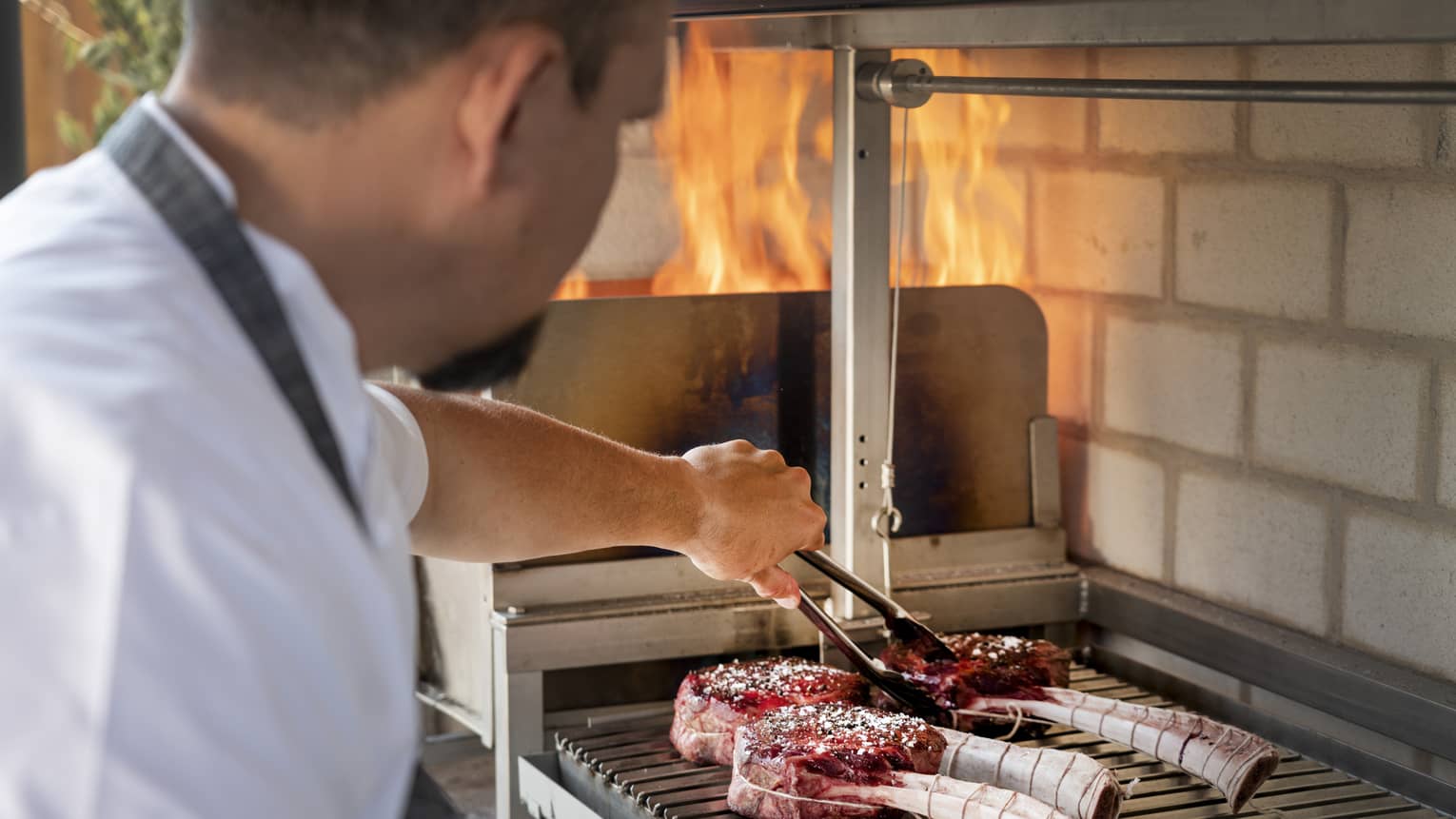 A man cooking large pieces of meat with bones on a grill.