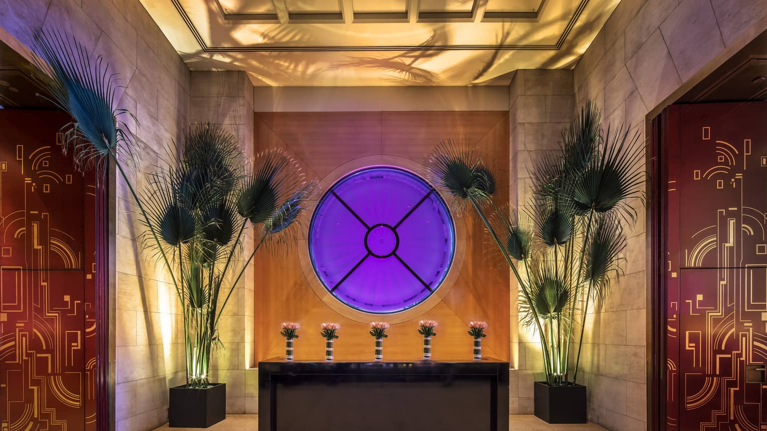 An ornate entryway to FIFTY with a circular window glowing purple, two tall green floral installations and red and gold abstract wall paper