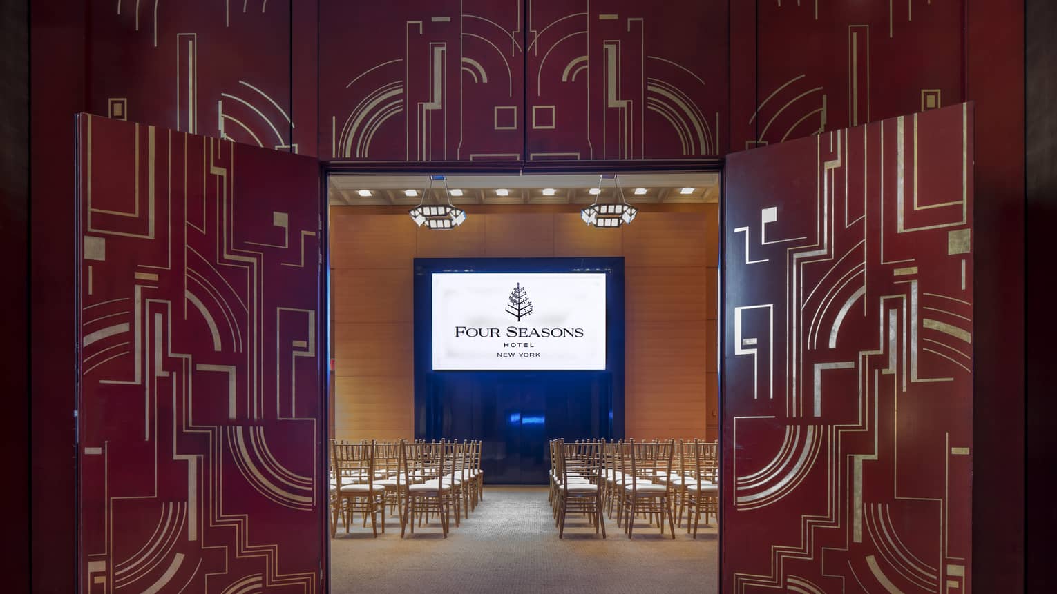 Two red and gold abstract and ornate doors are opened to reveal a board room with rows of chairs facing a screen with the Four Seasons logo on it 