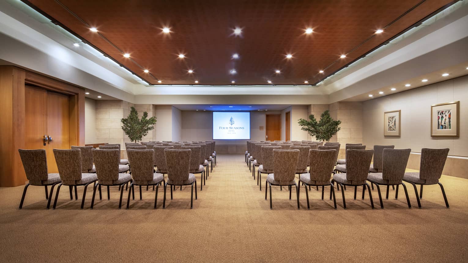 a wood-panelled function room with rows of chairs facing a presentation screen with two large green plants on either side of it 