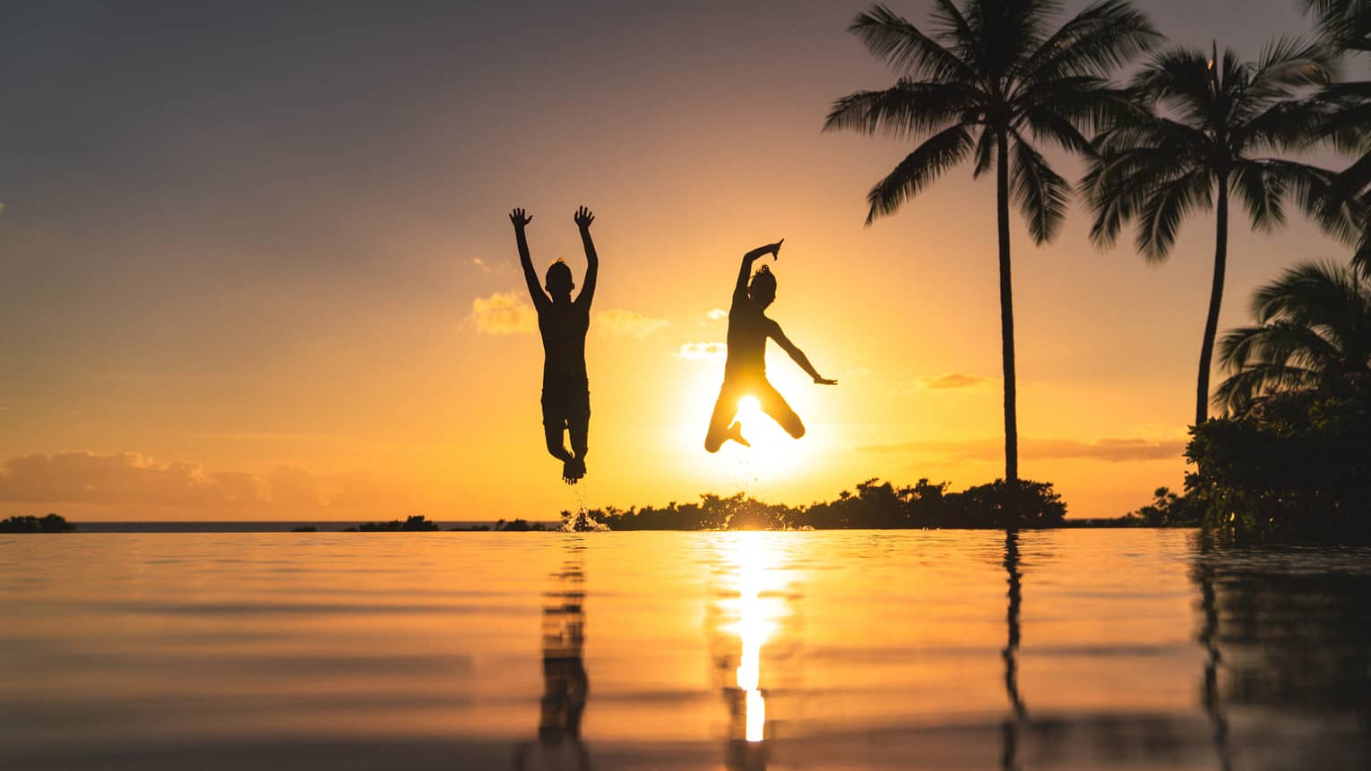 Silhouette of two children jumping above the water, sunset and palm trees in background 