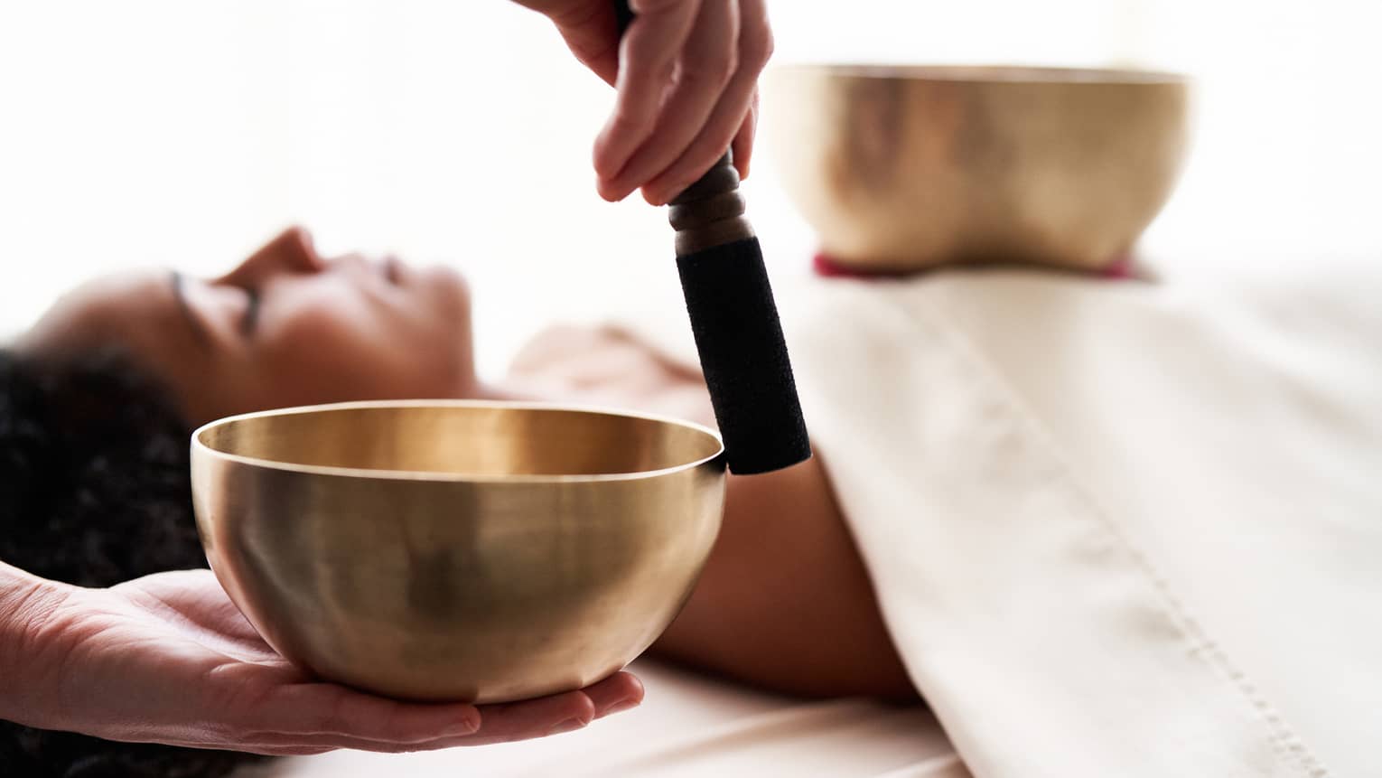 Close up of hands playing a singing bowl with a woman laying down on a spa treatment table in the background