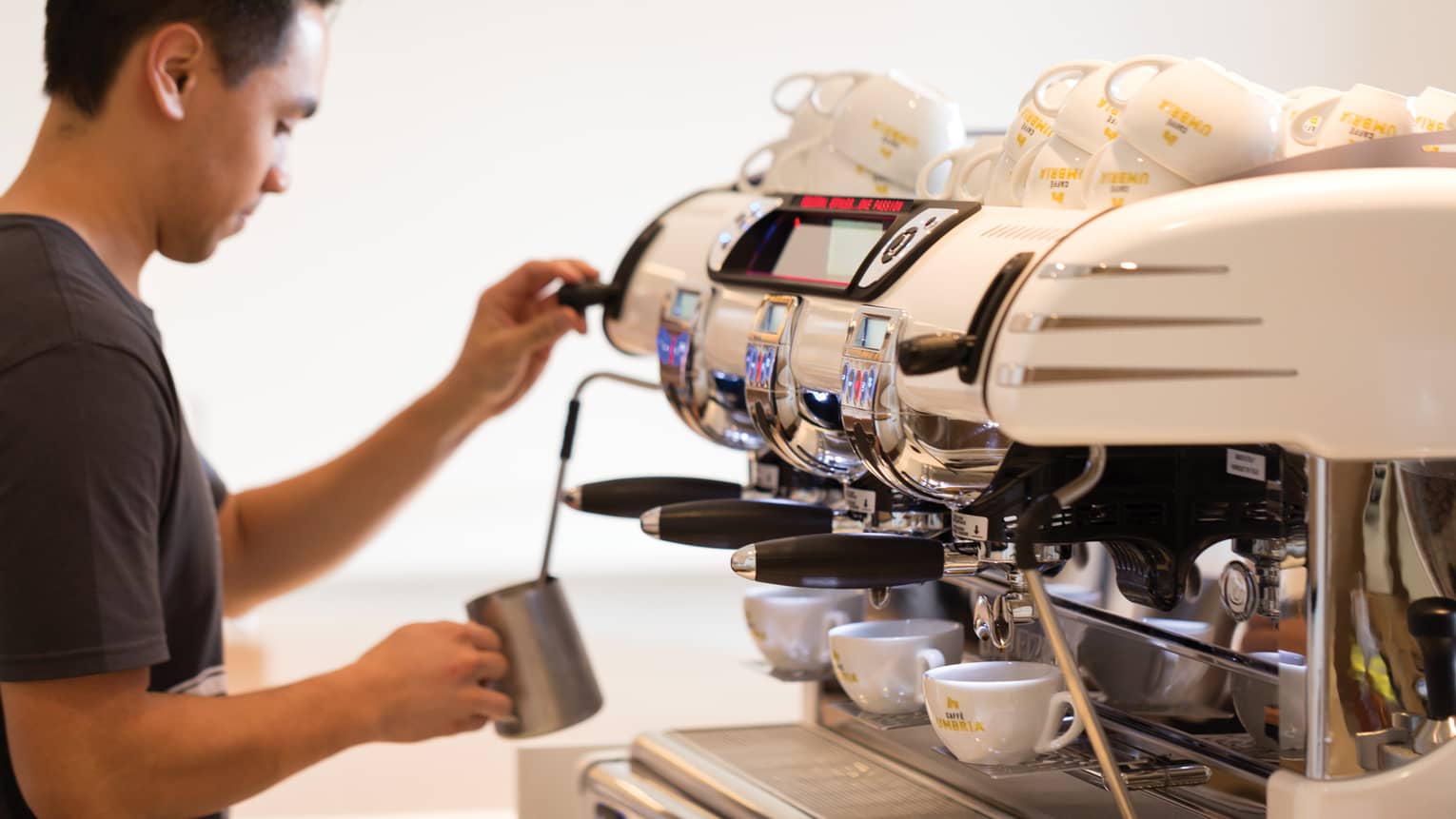 Barista stands in front of espresso machine lines with small white mugs, froths milk in silver jug