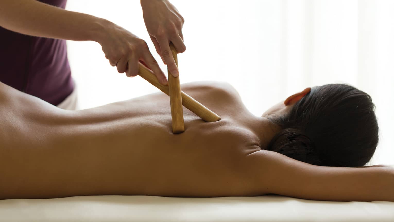 Masseuse presses two birchwood massage sticks into woman's bare back in spa