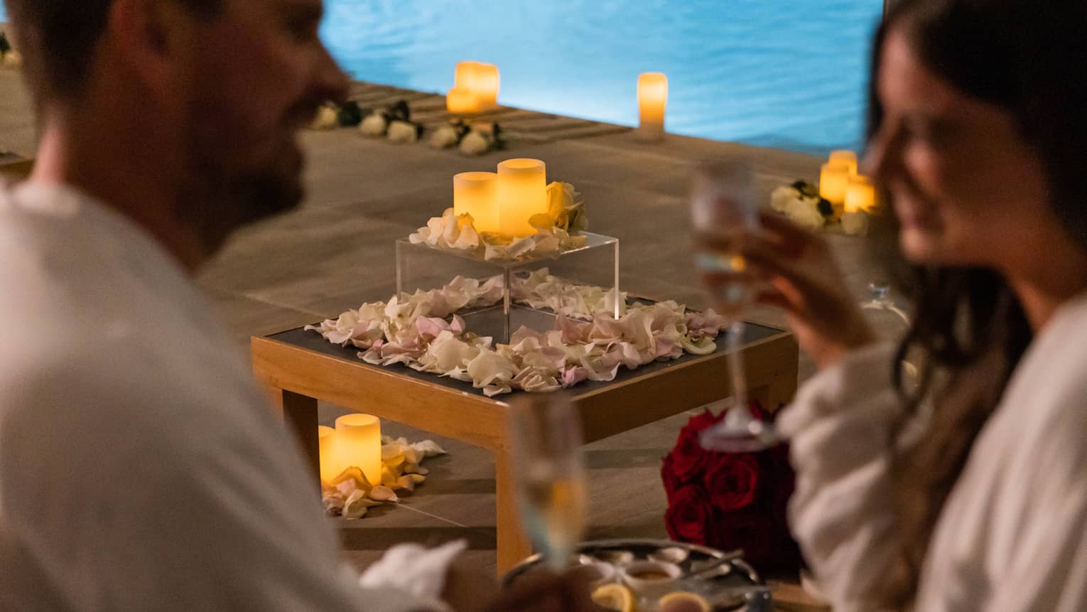 A couple with wine in hand near a pool surrounded by flowers petals and candles.