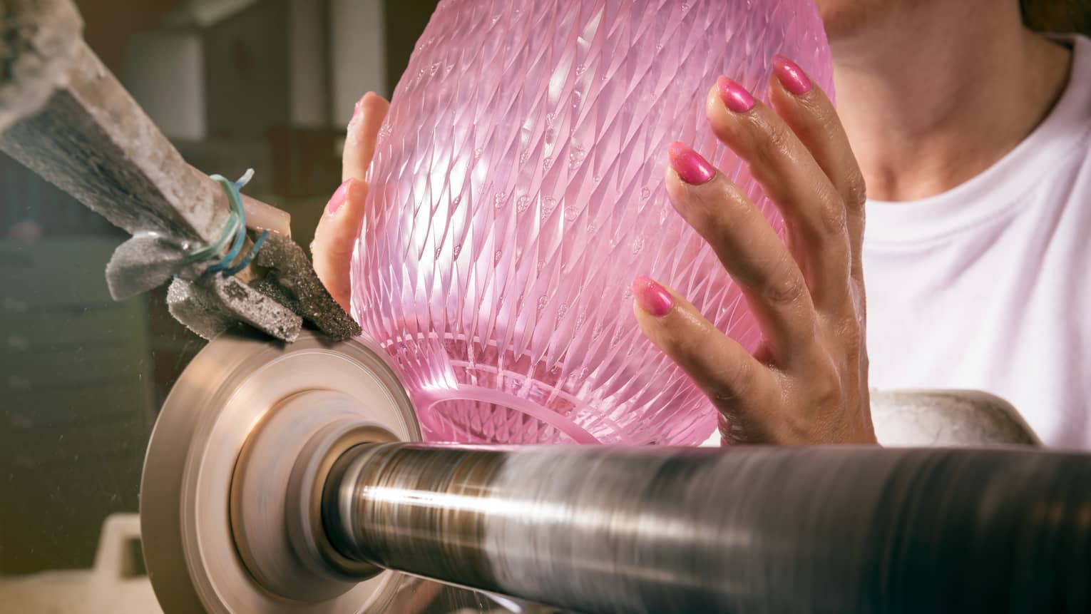 Hands with pink nails hold pink crystal vase atop glass-cutting machine