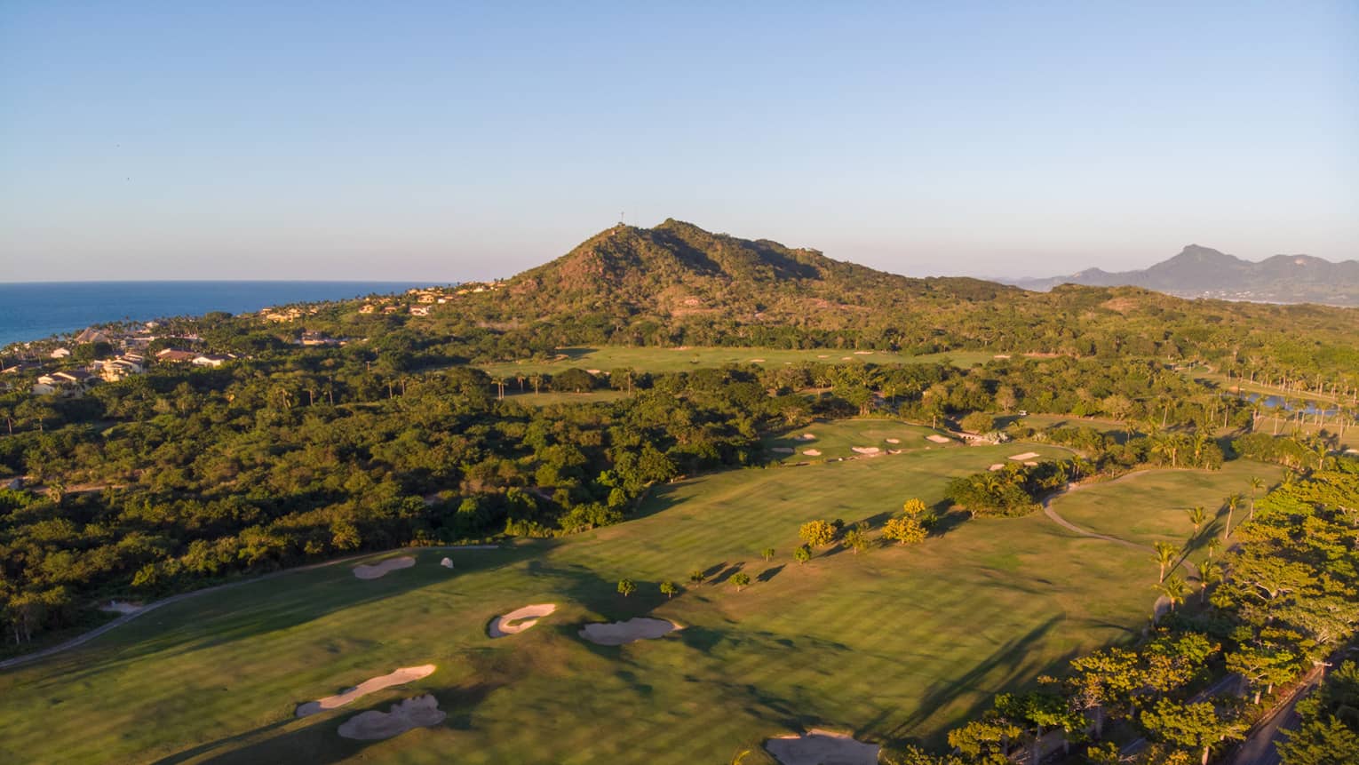 Aerial view of a golf course over the rolling coastal landscape; houses rising along the hillside, ocean in the distance.