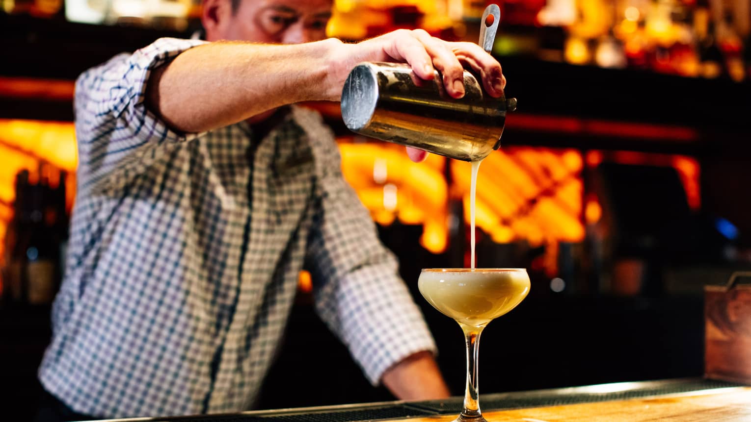 A man pouring a cocktail behind a wood bar.