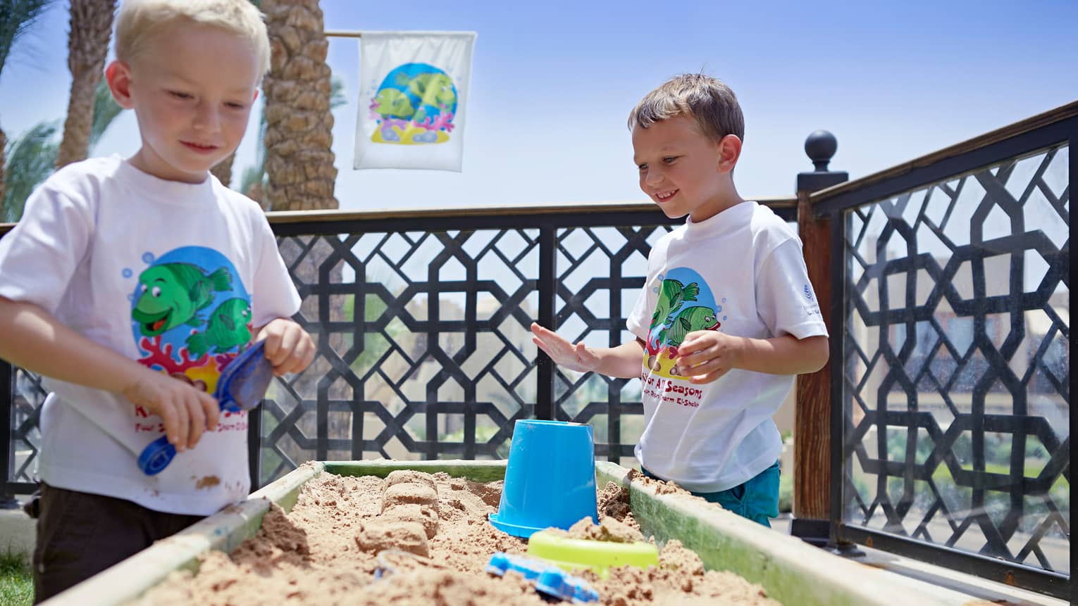 Two young boys in white T-shirts play at sand table with plastic buckets, shovels