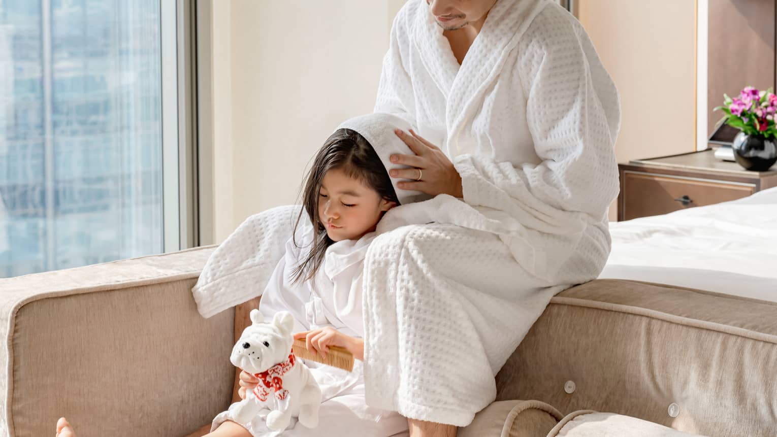 Mom and daughter in white robes sit on end-of-bed bench in guest room, girl holding stuffed dog as mom dries her hair with a towel