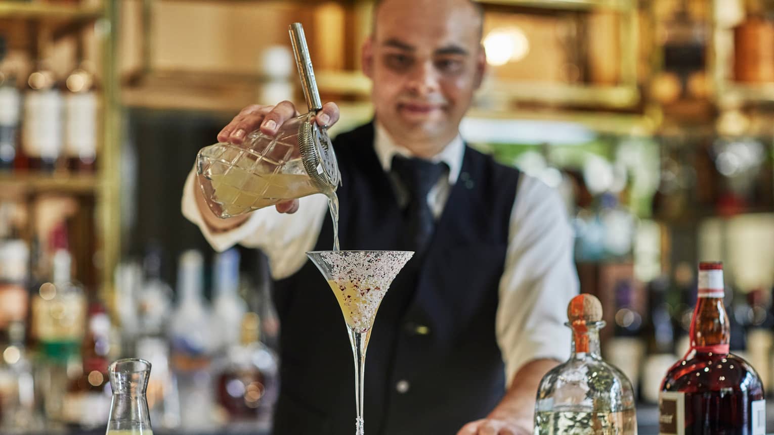 Bartender wearing black vest pours cocktail from crystal shaker into martini glass