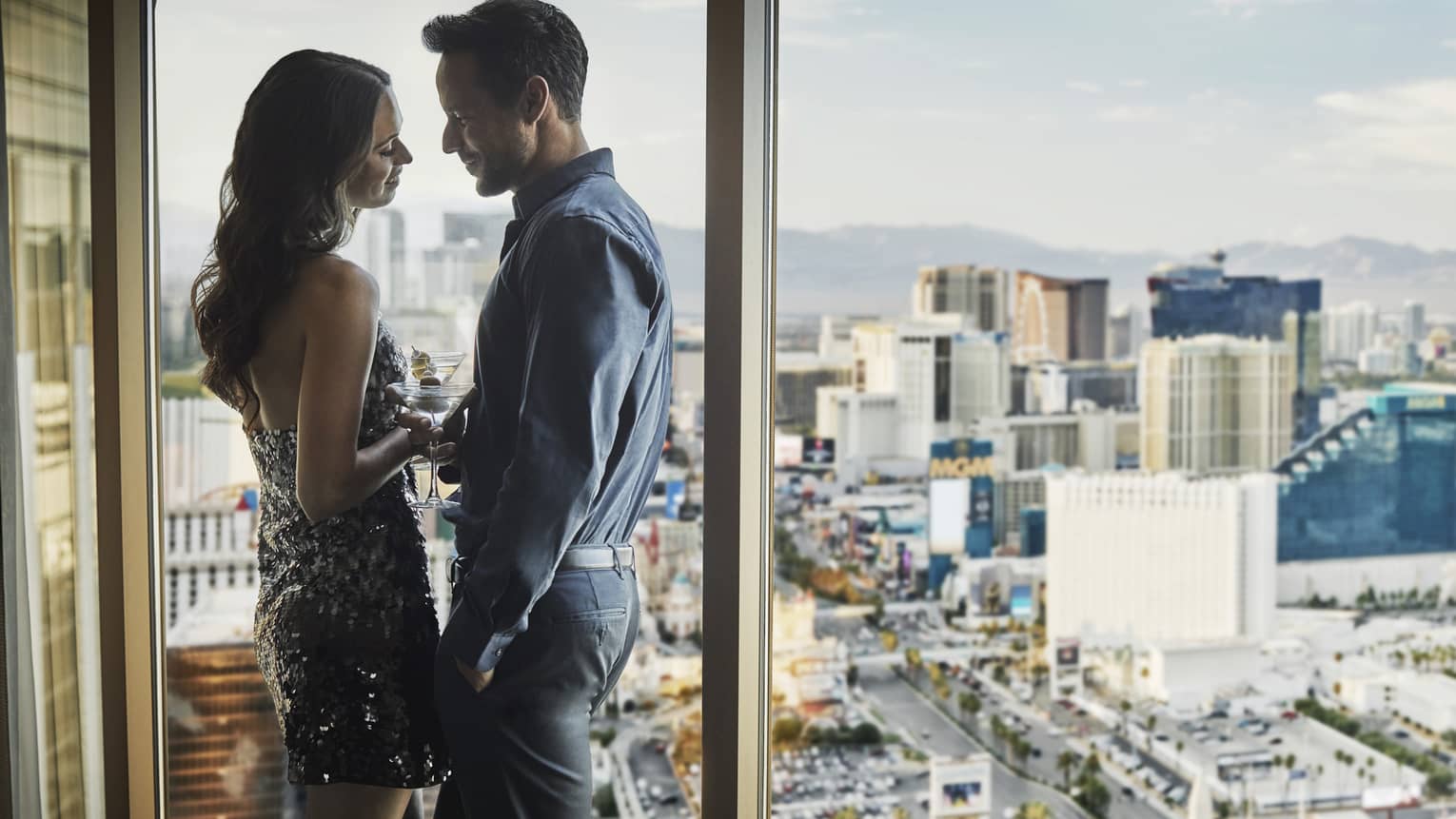 Couple holds cocktails, leans against floor to ceiling windows overlooking Las Vegas