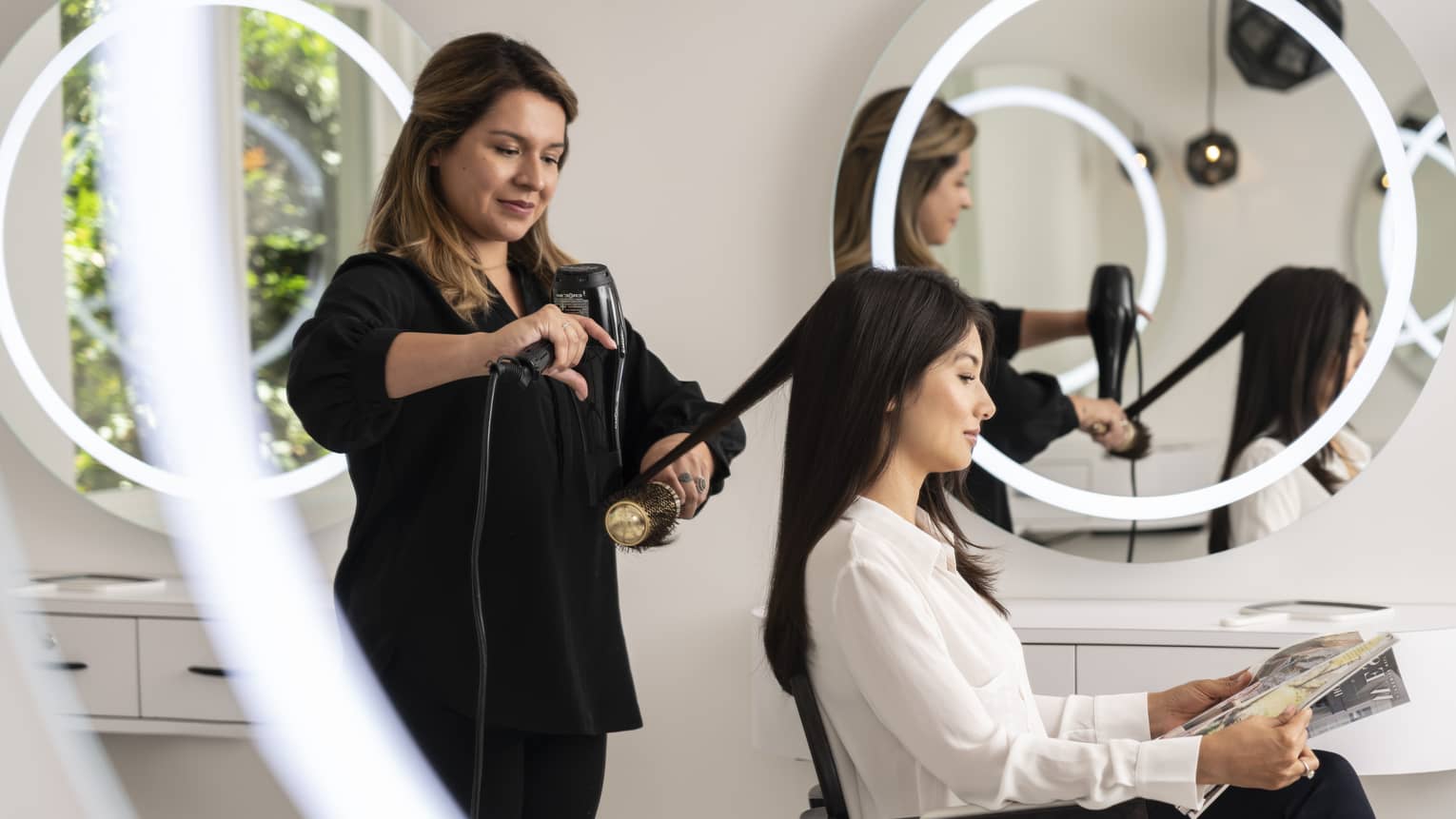A stylist blow drying a woman's hair while she sits at a salon.