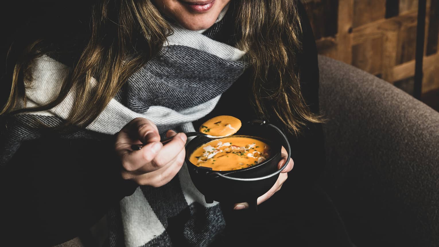 A woman with long light-brown hair and wearing a black jacket and grey-and-white scarf holds a cauldron-style bowl of orange-colored soup in one hand and a silver spoon in the other
