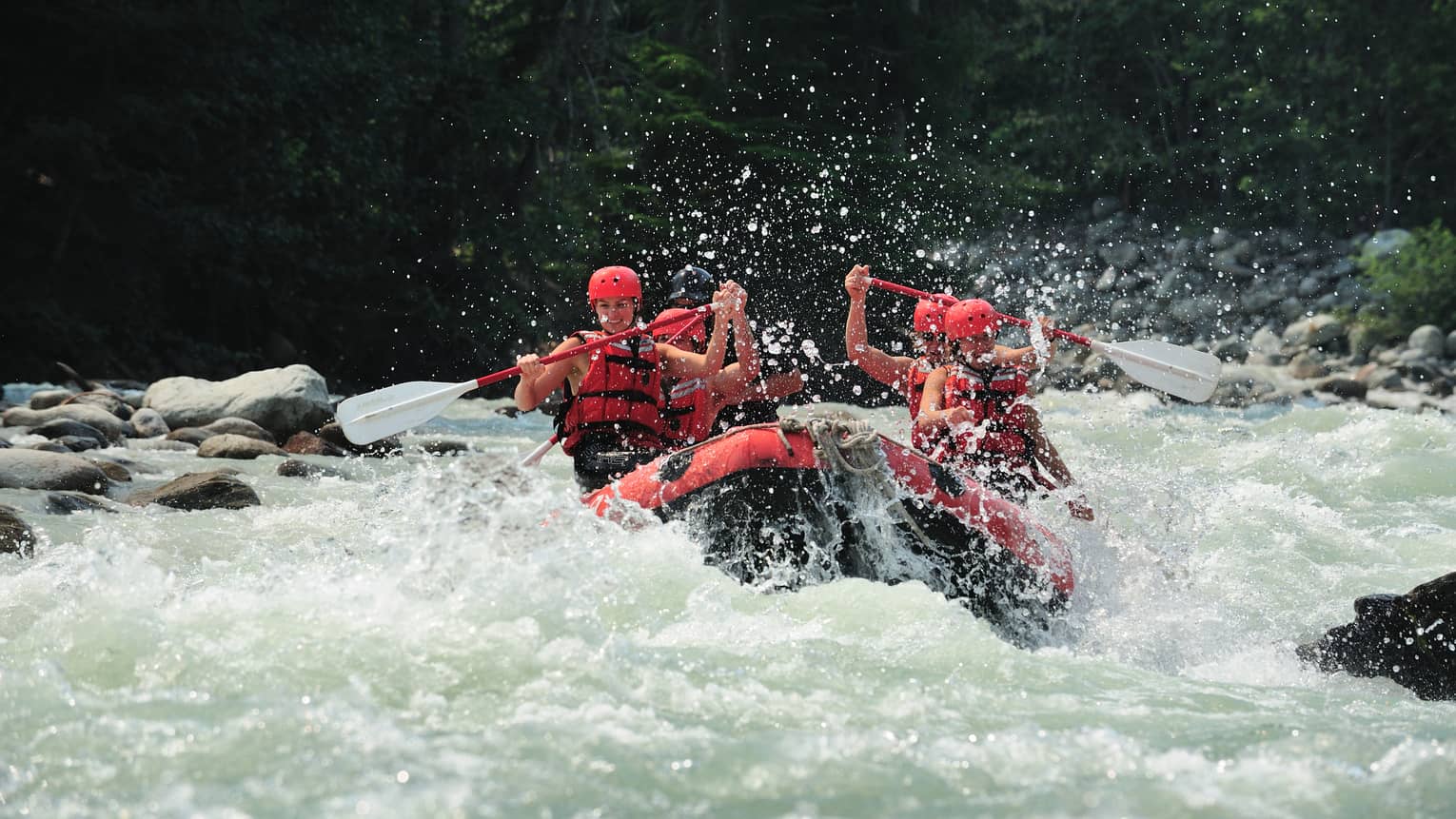 Smiling paddlers in red helmets and lifejackets steer an inflated raft through raging white water, passing evergreen trees.