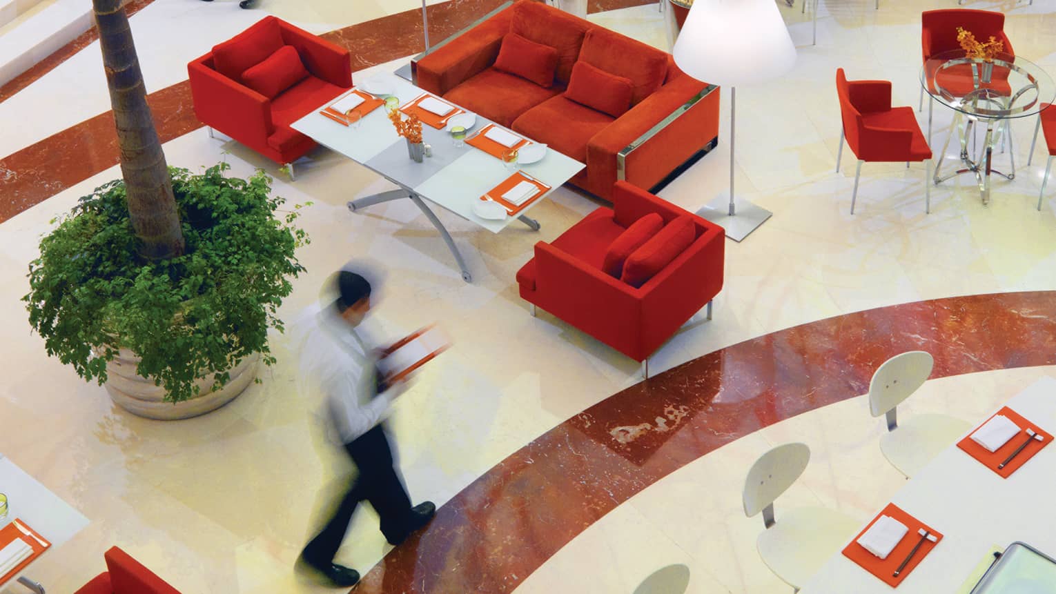 Aerial view of server walking quickly through Beymen Cafe, past red sofas and chairs and indoor tree