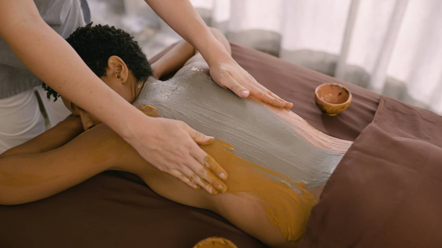 Hands rub clay across person's back as they lay on massage table