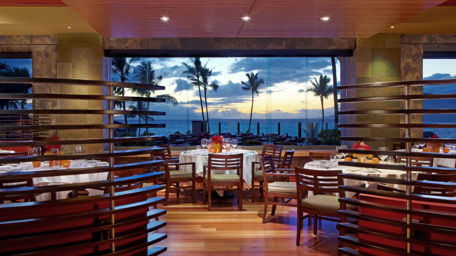 Spago dining room with wood decor, floor-to-ceiling windows looking out at palm trees, oceans and sunset