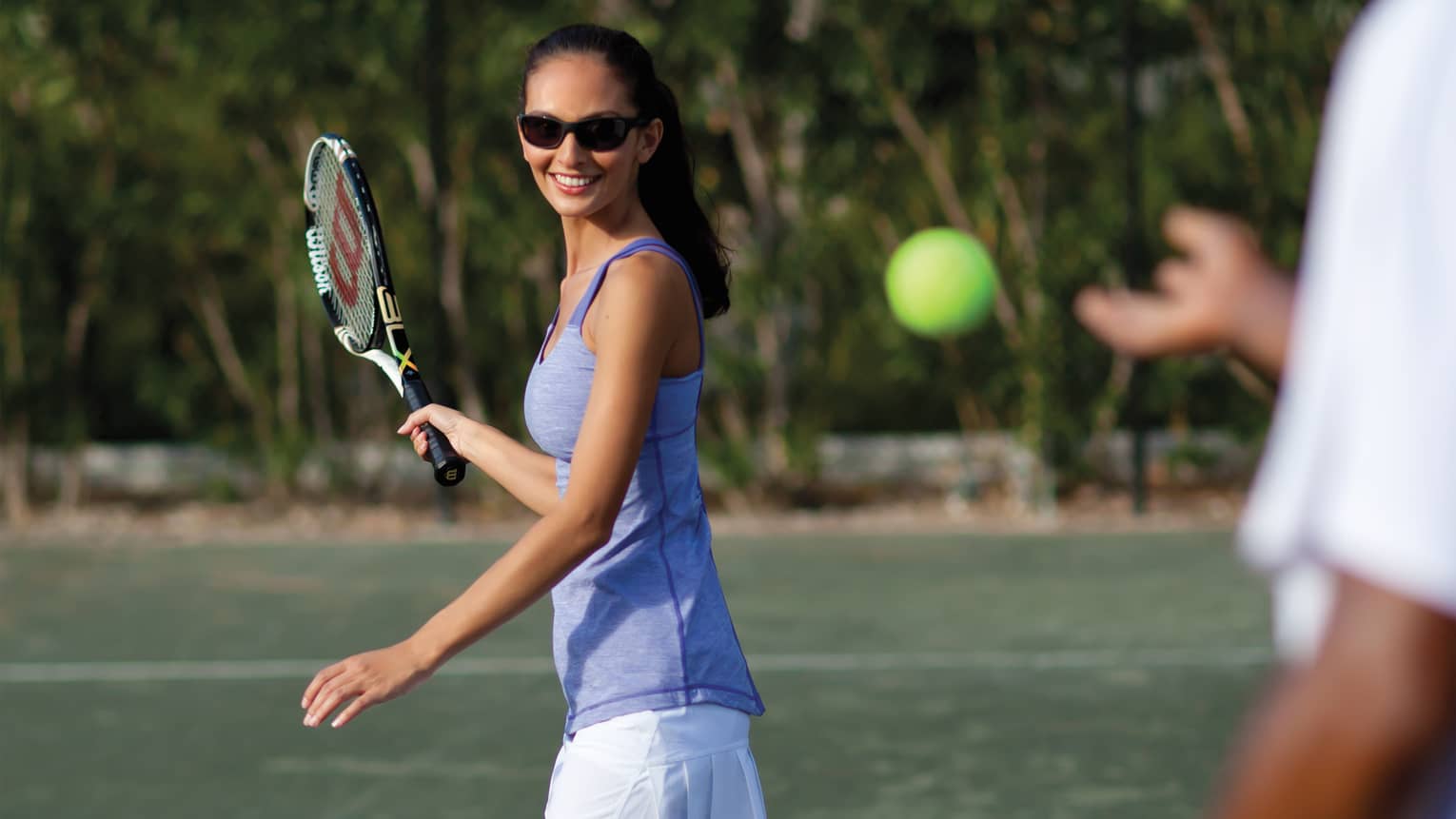 Woman in sunglasses and swings tennis racket as tennis ball approaches