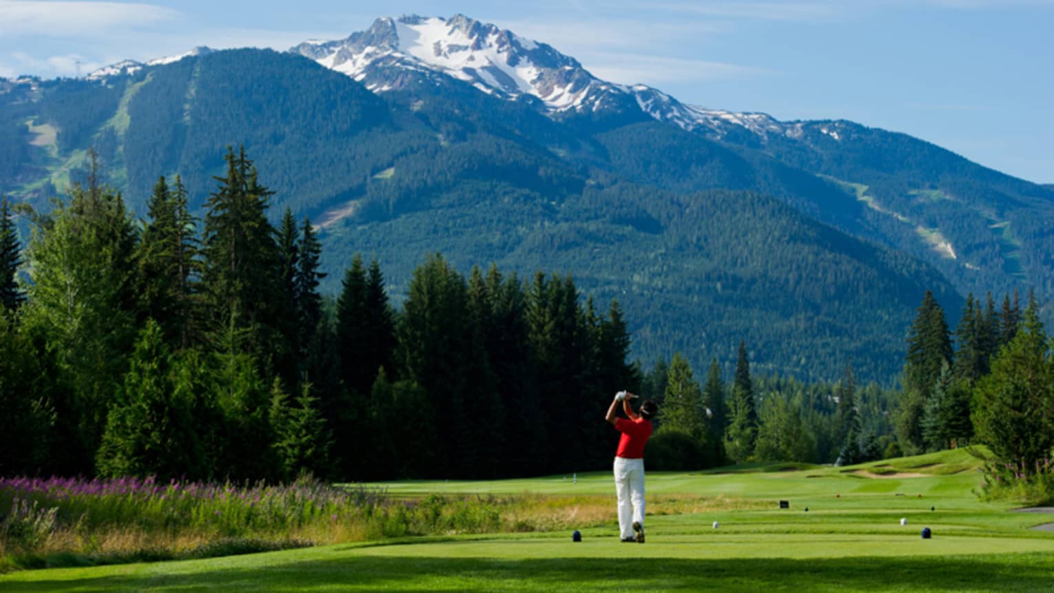 Man wearing red golf shirt swings club on green golf course surrounded by snow-capped mountains