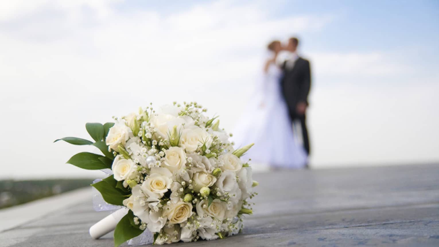White floral wedding bouquet on ground, bride and groom kissing in background