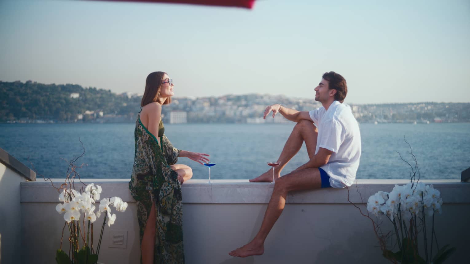 Couple sits facing each other on terrace wall, holding wine glasses, with Bosphorus water view