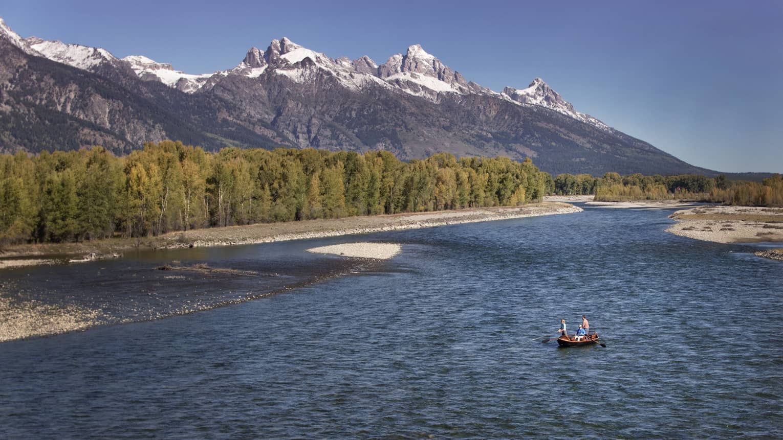 People fishing in boat on wide river by rocky shore, green trees, mountains