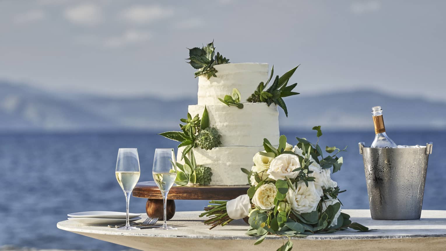 Three-tier wedding cake with green foliage decoration on outdoor table with matching bouquet, two glasses of champagne, bucket of champagne on ice