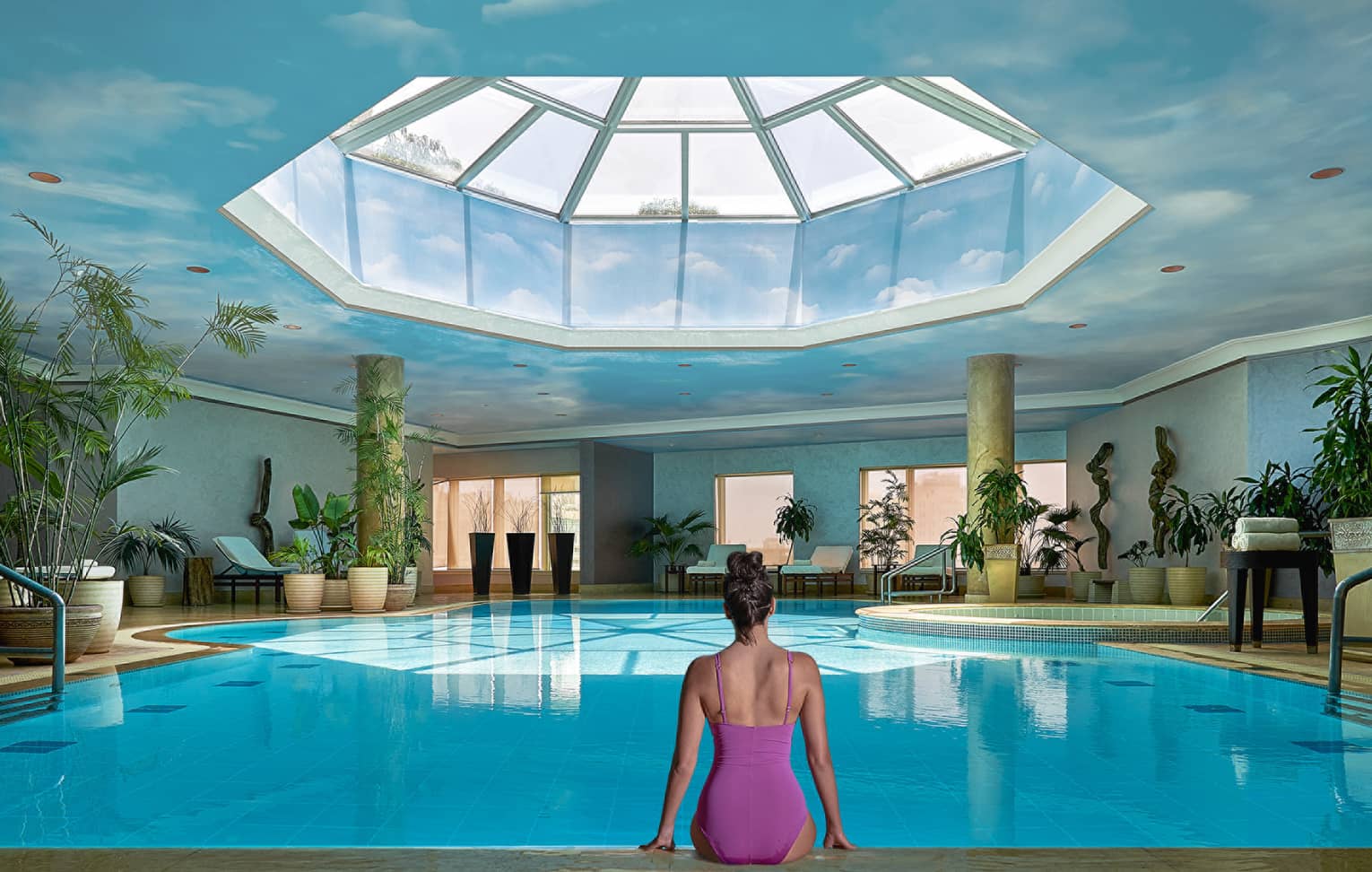 Woman in swimsuit seated on edge of indoor pool with blue ceiling and skylight above