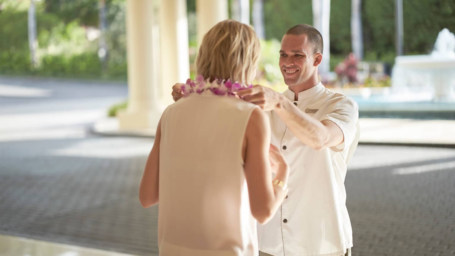 Man in white hotel uniform puts purple Hawaiian lei garland around woman's neck