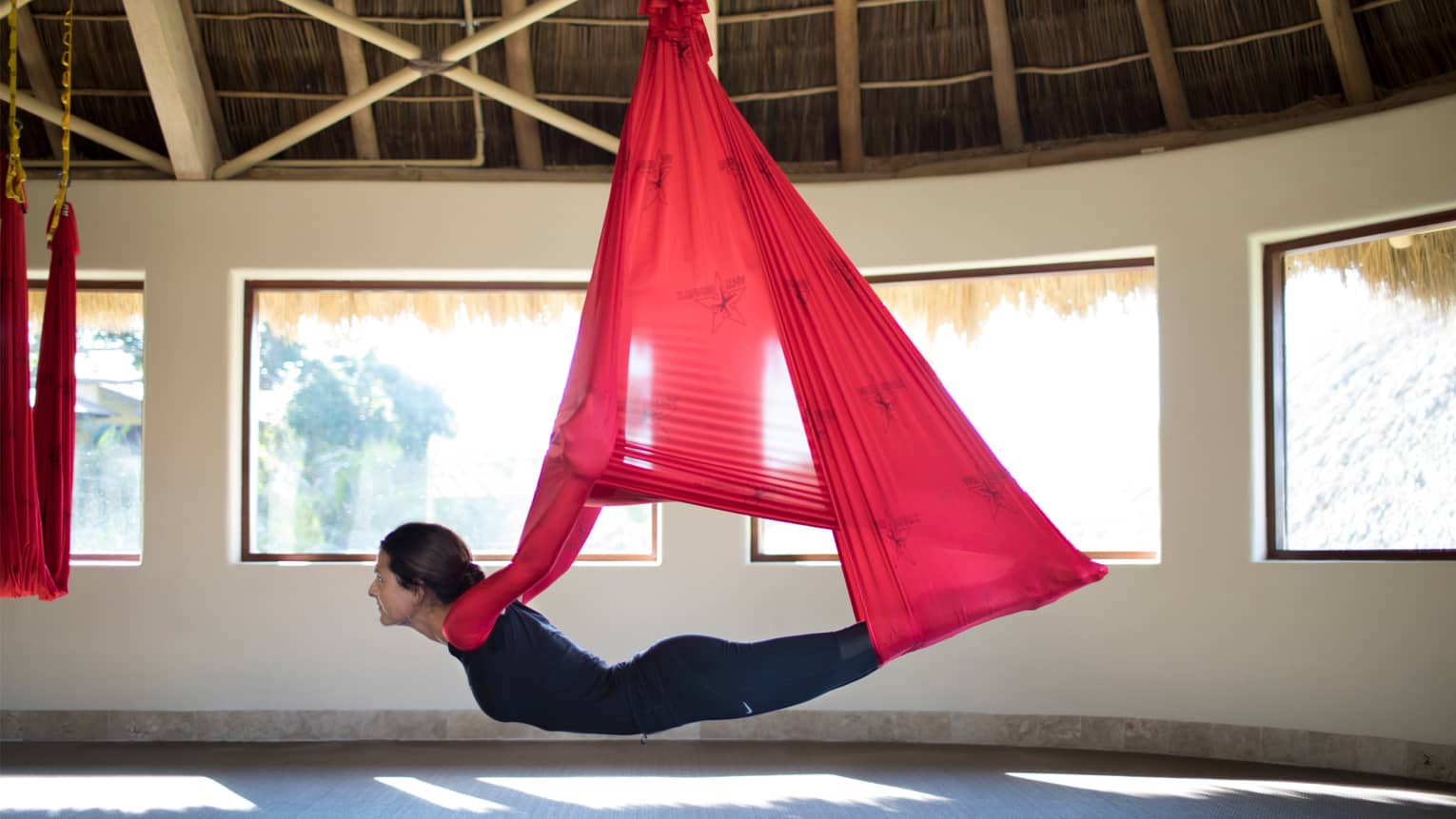 Woman hovers above ground, hanging from red fabric in anti-gravity yoga pose under thatched roof