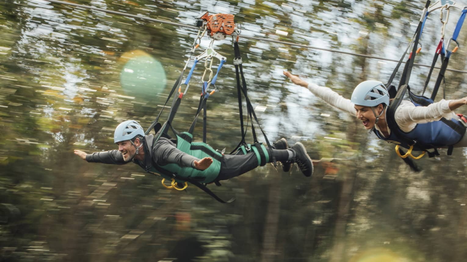 Close-up of a couple in full safety gear with wide smiles and arms outstretched as they zip-line past a blur of treetops.